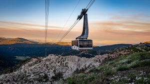 A cable car labeled "Palisades Tahoe" ascends over rugged mountainous terrain. The backdrop features distant peaks, partly obscured by clouds during sunset. The scene is painted in shades of green from the trees, blue from the sky, and orange from the setting sun. It resembles views you might see on a ride in a Mammoth tram.