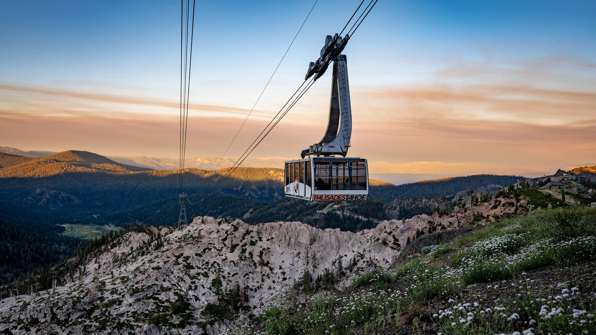 A cable car labeled "Palisades Tahoe" ascends over rugged mountainous terrain. The backdrop features distant peaks, partly obscured by clouds during sunset. The scene is painted in shades of green from the trees, blue from the sky, and orange from the setting sun. It resembles views you might see on a ride in a Mammoth tram.