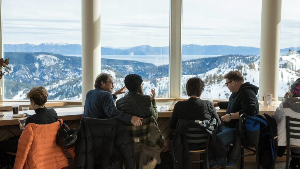 A group of people and dogs sitting at a table overlooking snowy mountains at Palisades Tahoe.