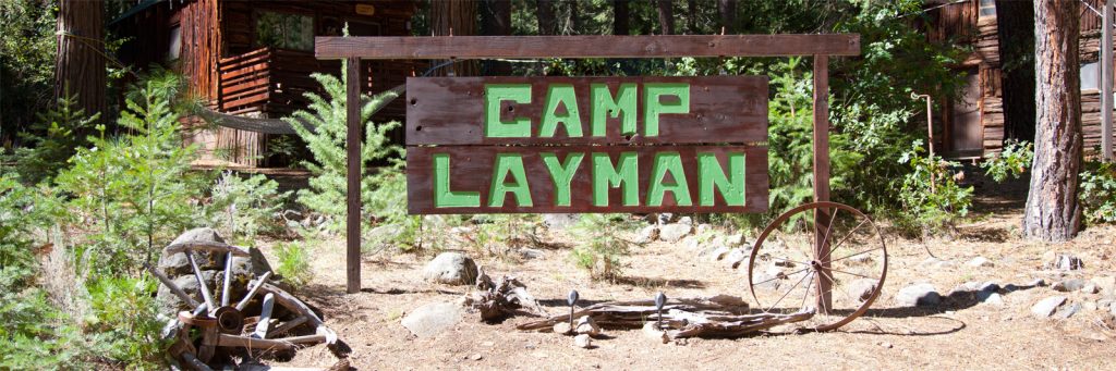 A wooden sign with "Camp Layman" in large green letters stands outside, held up by two wooden posts. It is surrounded by trees, bushes, an old wagon wheel, and a log cabin. The scene gives off a wilderness camp vibe in the High Sierra region.