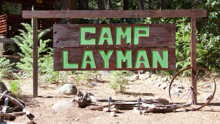 A wooden sign with "Camp Layman" in large green letters stands outside, held up by two wooden posts. It is surrounded by trees, bushes, an old wagon wheel, and a log cabin. The scene gives off a wilderness camp vibe in the High Sierra region.