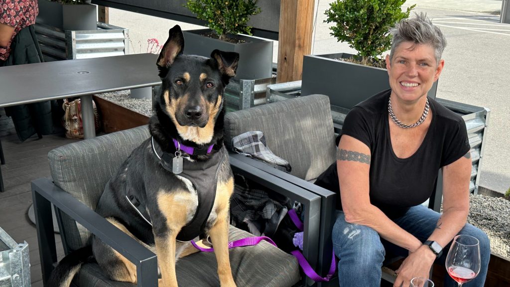 A person with short gray hair sits at an outdoor café table in Healdsburg, dressed in a black shirt and jeans. Beside them, a black and tan dog relaxes on its own chair. A wine glass sits among other items on the table. The background is filled with lush greenery, creating a serene atmosphere that feels refreshing and revitalizing.