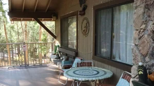 At Wawona Cabin, you'll find a quaint wooden porch with a small round table. The table is covered with an intricately crocheted tablecloth and surrounded by three comfortable chairs, perfect for sitting and relaxing with your dog. A stone wall backdrops the setup, accentuated by a simple "Welcome" sign that invites you in. Tall trees and lush greenery envelop Cabin 28, providing a tranquil setting ideal for spending quiet moments outside with your furry companion.