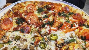Engfer's pizza offers a unique experience with its two contrasting halves. One side is adorned with ripe tomatoes, creamy feta cheese, and fragrant pesto. The other supports a savory mix of earthy mushrooms, spicy jalapeños, juicy sausage, and fresh spinach. Each half maintains its own character while sharing the same crisp crust. The pizza arrives on a metal tray that highlights the vibrant colors and textures of this carefully crafted dish. Perfect for enjoying with your furry friend in tow.