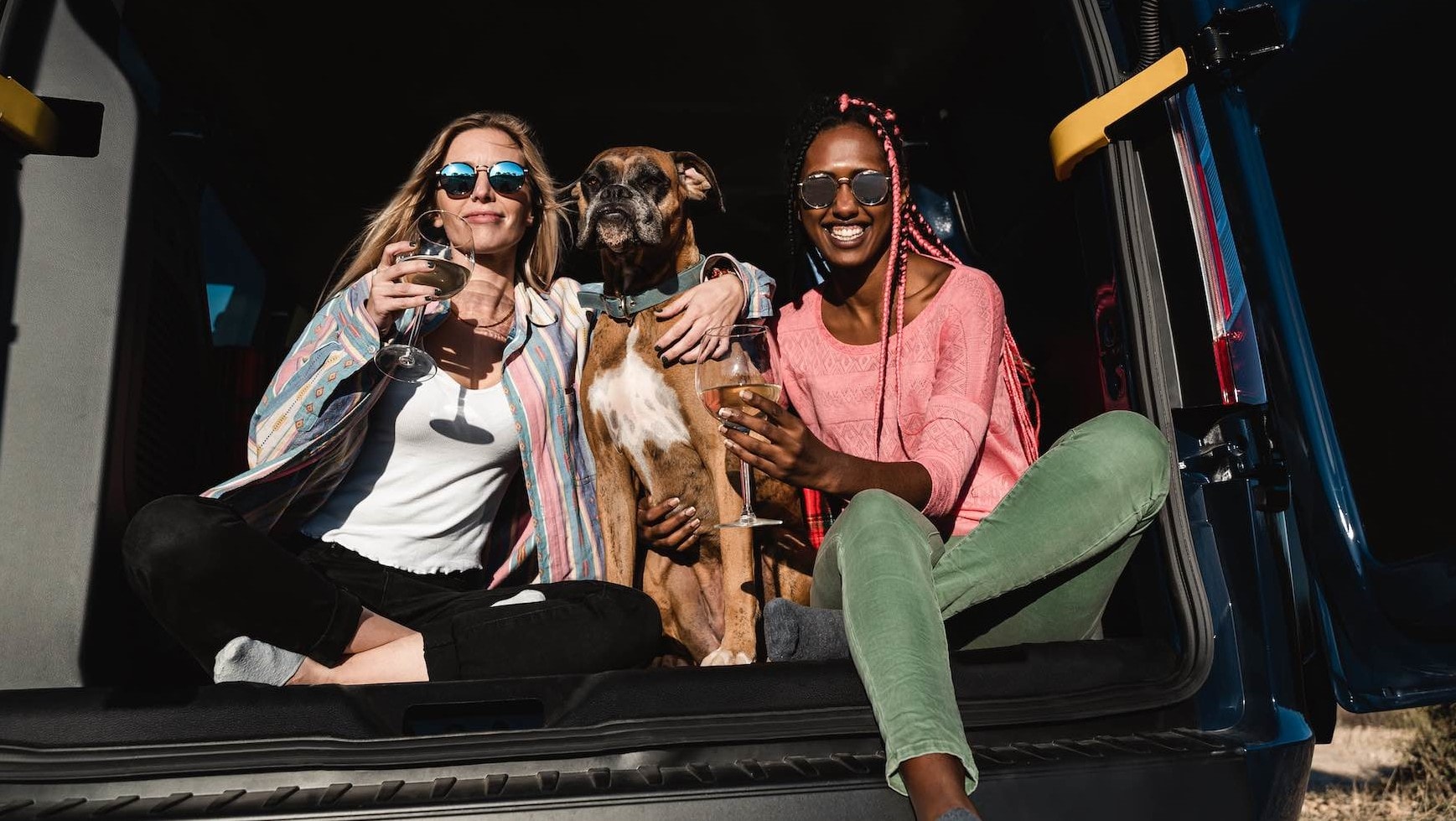 Two women with dog celebrating with wine together in camper van.