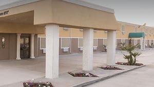 A vibrant dog park bathed in sunlight features abundant greenery with mature trees, offering ample shade for leisurely outdoor activities. In the distance is a well-equipped playground that presents an additional activity area. An inviting picnic table spreads out nearby, ideal for enjoying meals al fresco or taking relaxing breaks from play. A neatly-placed trash bin aids in maintaining the park's cleanliness for everyone's enjoyment. Dominating the scene is a generously wide dirt path gracefully curving through the expanse of grass, promising exploratory walks and lively fetch games.