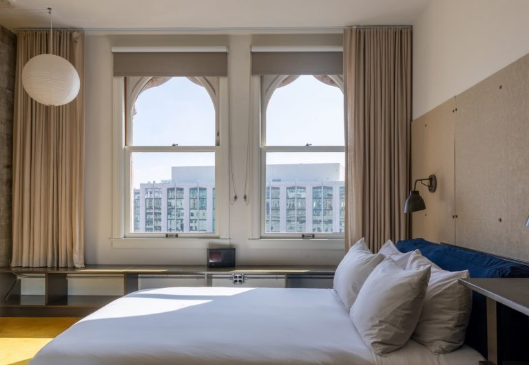 Dog-friendly Kasa hotel room in Downtown LA with large windows, beige curtains, built-in shelves, bed, and city views.