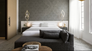 A modern bedroom at La Peer Hotel includes a platform bed with white linens, two angular pendant lights suspended above wooden side tables, and a compact gray sofa positioned at the end of the bed. Large windows, dressed with sheer white curtains, allow sunlight to fill the space.