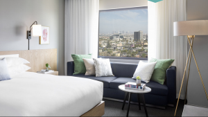 A Hollywood hotel room with a neatly made bed sits adjacent to a dark blue sofa positioned beside the window. The window offers views of the city streets below. Next to the sofa, a round side table holds books and a small green plant. A floor lamp stands nearby for evening light. Above the wooden headboard, framed wall art completes the space.