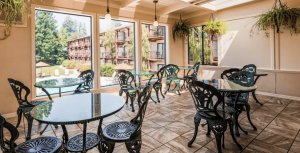 A vibrant dog park bathed in sunlight features abundant greenery with mature trees, offering ample shade for leisurely outdoor activities. In the distance is a well-equipped playground that presents an additional activity area. An inviting picnic table spreads out nearby, ideal for enjoying meals al fresco or taking relaxing breaks from play. A neatly-placed trash bin aids in maintaining the park's cleanliness for everyone's enjoyment. Dominating the scene is a generously wide dirt path gracefully curving through the expanse of grass, promising exploratory walks and lively fetch games.