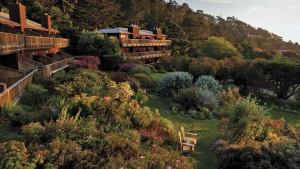 Stanford Inn offers a landscape of wooden buildings set against a rocky hillside. The area features diverse greenery, colorful flowers, and trees. A wooden bench provides a vantage point for the scene under a clear sky.