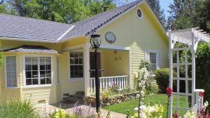 This single-story, yellow house welcomes dogs and their owners. White trim frames the windows and doors. The front porch offers space to relax after a walk, while a bay window provides views of the landscaped garden. A black lamp post stands close to the main entrance, lighting the path for evening strolls. On the right side of the yard, a white garden arbor marks an entryway—ideal for games of fetch or exploring with your dog in tow.