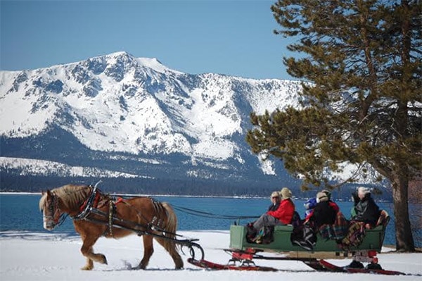 Horse pulls sled-style buggy filled with people over snow covered meadow with snow covered mountains in distance