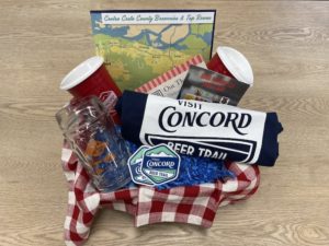 A vibrant dog park bathed in sunlight features abundant greenery with mature trees, offering ample shade for leisurely outdoor activities. In the distance is a well-equipped playground that presents an additional activity area. An inviting picnic table spreads out nearby, ideal for enjoying meals al fresco or taking relaxing breaks from play. A neatly-placed trash bin aids in maintaining the park's cleanliness for everyone's enjoyment. Dominating the scene is a generously wide dirt path gracefully curving through the expanse of grass, promising exploratory walks and lively fetch games.