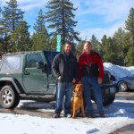 2 men and a dog stand in snowy parking lot