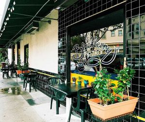 A vibrant dog park bathed in sunlight features abundant greenery with mature trees, offering ample shade for leisurely outdoor activities. In the distance is a well-equipped playground that presents an additional activity area. An inviting picnic table spreads out nearby, ideal for enjoying meals al fresco or taking relaxing breaks from play. A neatly-placed trash bin aids in maintaining the park's cleanliness for everyone's enjoyment. Dominating the scene is a generously wide dirt path gracefully curving through the expanse of grass, promising exploratory walks and lively fetch games.
