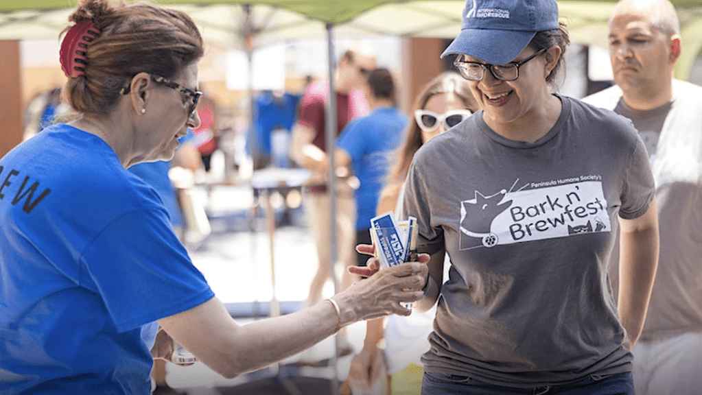 At the Bark n' Brewfest, a woman is cheerfully handing a drink to another woman amidst the lively outdoor event. - Dogtrekker