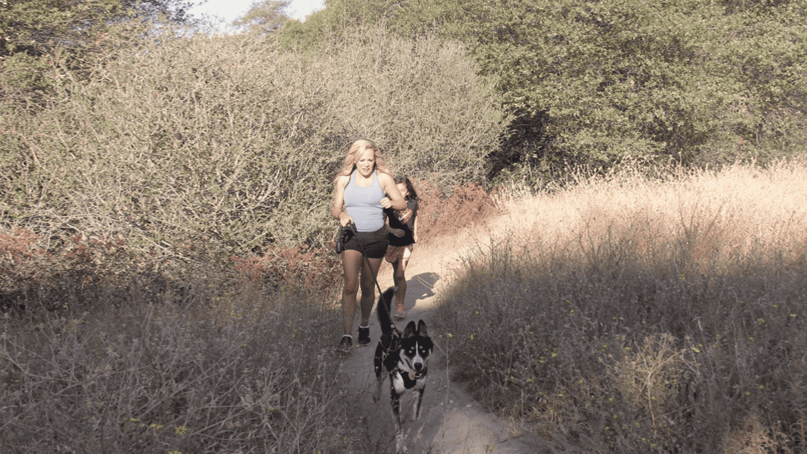 A woman and girl jogging a dog down a dirt trail.