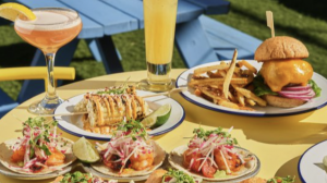 A table features plates of shrimp tacos garnished with sliced radishes and herbs, street corn covered in cheese, and a cheeseburger paired with fries. Drinks include a pink cocktail and a tall glass filled with a yellow beverage. In the background, blue picnic benches offer seating where dogs are welcome.