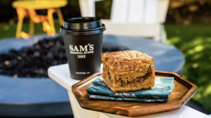 A piece of coffee cake rests on a wooden plate beside a green napkin and a black cup marked "Sam's General Store" on an outdoor table. Chairs and leafy plants surround the area, creating a setting where dogs are welcome to relax alongside their owners.