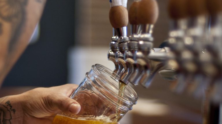 A vibrant dog park bathed in sunlight features abundant greenery with mature trees, offering ample shade for leisurely outdoor activities. In the distance is a well-equipped playground that presents an additional activity area. An inviting picnic table spreads out nearby, ideal for enjoying meals al fresco or taking relaxing breaks from play. A neatly-placed trash bin aids in maintaining the park's cleanliness for everyone's enjoyment. Dominating the scene is a generously wide dirt path gracefully curving through the expanse of grass, promising exploratory walks and lively fetch games.