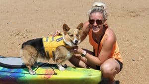 A corgi wearing a yellow life jacket stands on top of a kayak with a smiling woman on the beach of Lake Tahoe.