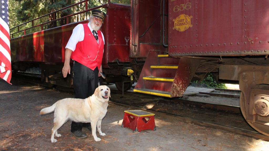 A conductor stands alongside a locomotive where a yellow lab is waiting to board the train.