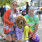 A man and a woman, both dressed in brightly colored bohemian clothing, kneel beside their large, fluffy dog. The dog is also wearing a vibrant outfit. They are outdoors at Woofstock, surrounded by people and tents. The man holds a sign that reads "Free Tibet.