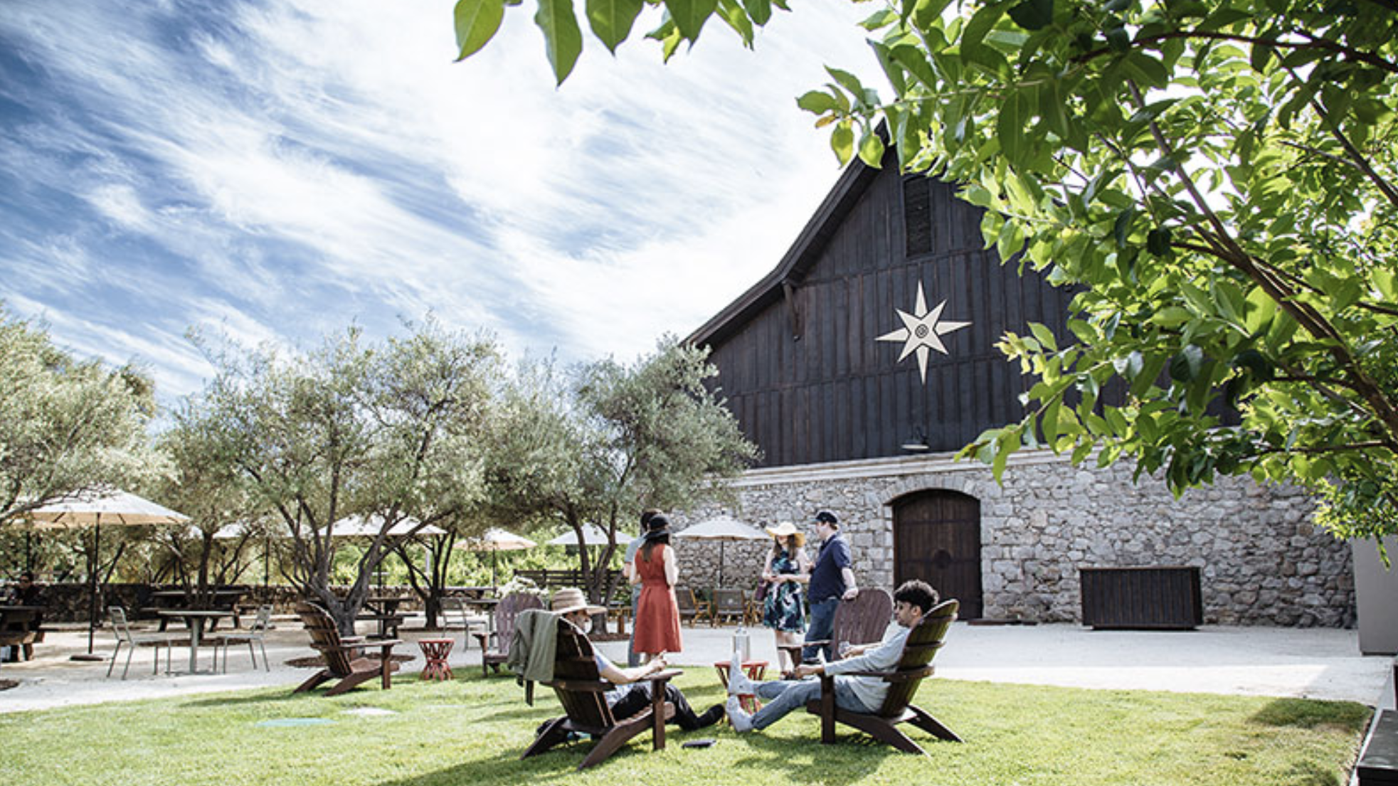 Individuals relish the warmth of the sun at open-air seating adjacent to an expansive, charming barn adorned with a star accent.
