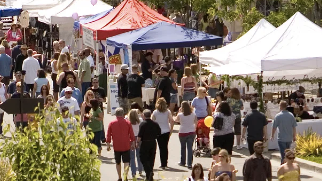In Downtown Livermore, an outdoor market unfolds with rows of canopy tents lining the street. Shoppers walk among the stalls, passing under clear skies. Many carry bags filled with their purchases; some hold balloons. Vendors offer an array of goods for sale. The scene is complemented by surrounding trees and greenery.