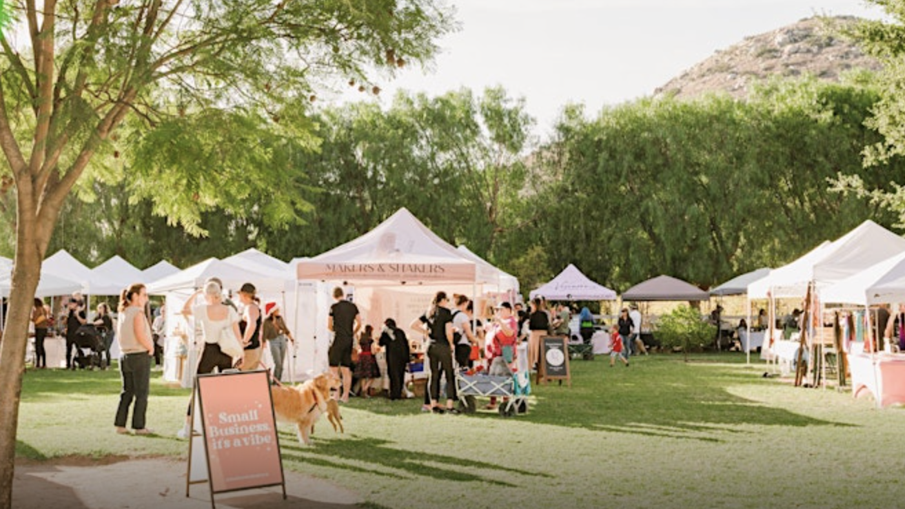 On a sunny summer day, a community outdoor market is alive with activity as people browse vendor tents. A sign nearby reads "Small Business Saturday". Among the visitors, a person walks their dog on a leash. The stall-lined paths are set against green trees and hilly terrain.