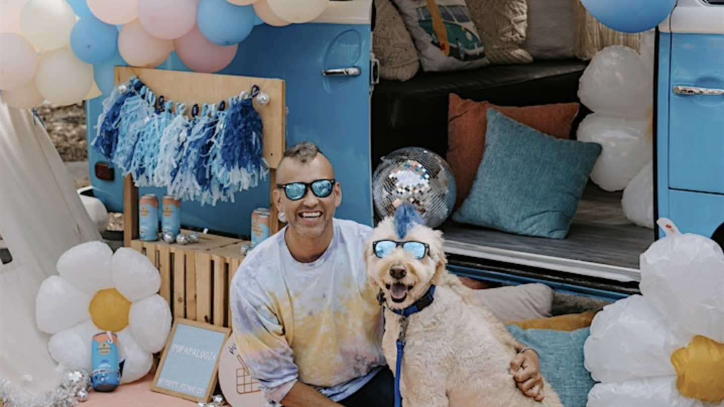 A man and his light-furred dog, both donning sunglasses, sit by a blue van at Pupapalooza. The area is decorated with balloons, blue and white tassels, flowers, and a disco ball. The man smiles at the camera as his dog sits contentedly next to him.