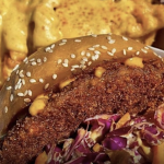 A close-up shows a crispy fried chicken sandwich topped with shredded cabbage and sauce in a sesame seed bun. Behind the sandwich, crinkle-cut fries drizzled with melted cheese sauce sit in a takeout container from San Clemente Market.