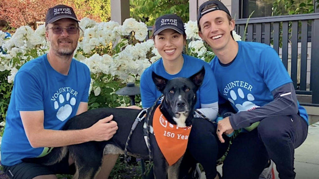 Three volunteers in blue shirts and hats stand with a black dog in front of a garden filled with white flowers. The dog from the East Bay SPCA wears an orange bandana that says "150 Years.