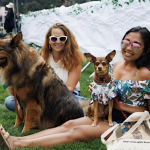 At the Bark at the Park event, two women sit on the grass with their dogs. The woman on the left, wearing sunglasses, is next to a large fluffy dog. The woman on the right, also in sunglasses, has a small dog in her lap. Both dogs wear bandanas and sit against a white tarp backdrop.