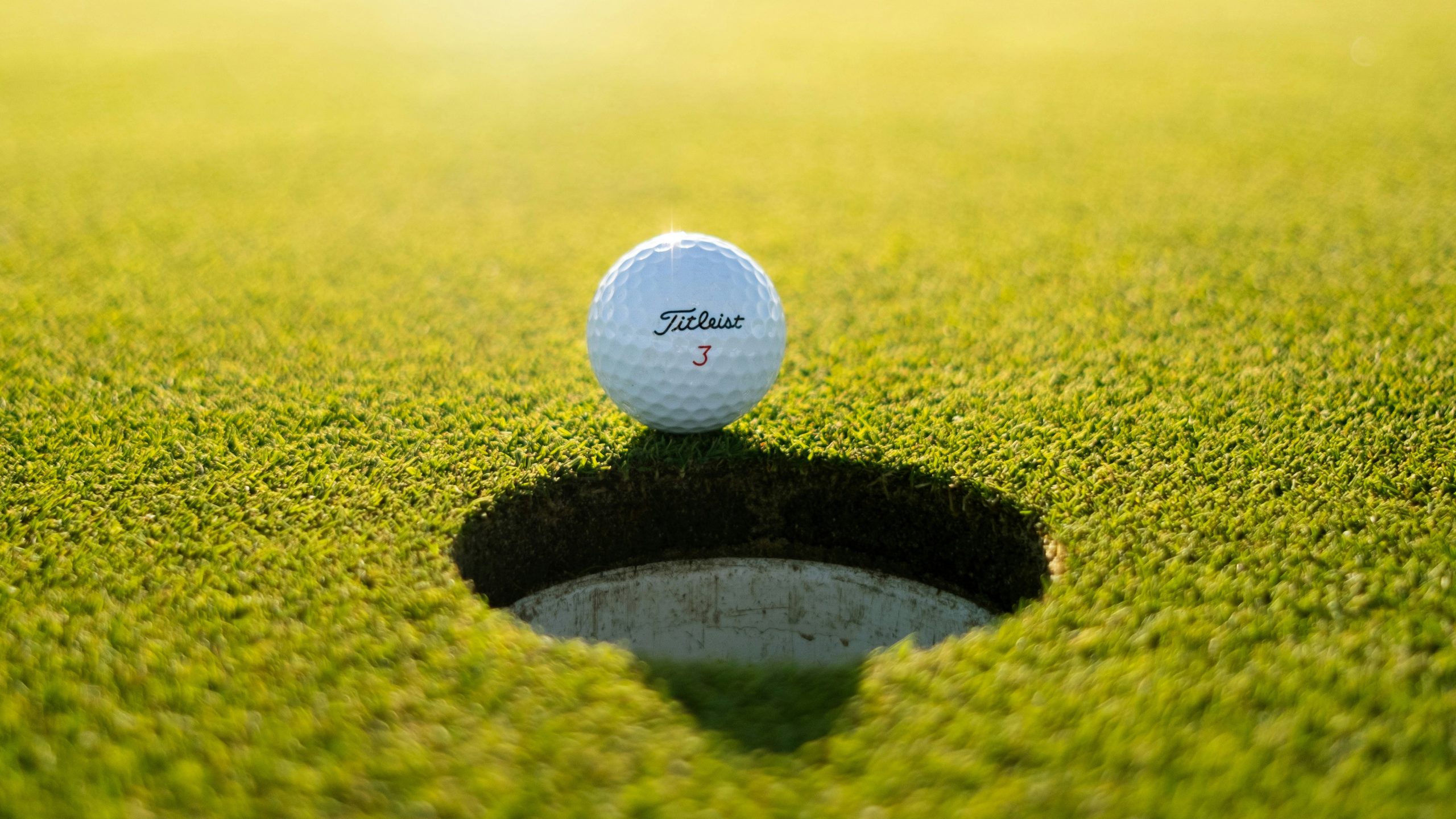 A Titleist 3 golf ball balances precariously on the lip of a hole during an Invitational game, bathed by shimmering sunlight on a verdant, well-manicured golf turf.