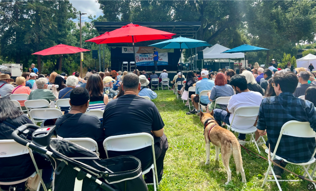 People sit in white folding chairs, facing an outdoor stage situated under tree cover. Red, blue, and teal umbrellas offer shade from the sun. Some attendees have brought strollers and dogs. A banner on the stage reads "Welcome," though some of the text is obscured. The event is popular among dog owners and a highlight of the summer season.Explore this Event:1. Find shaded seating under colorful umbrellas.2. Bring your dog to enjoy the pet-friendly atmosphere.3. Experience one of summer's key gatherings for dog enthusiasts.