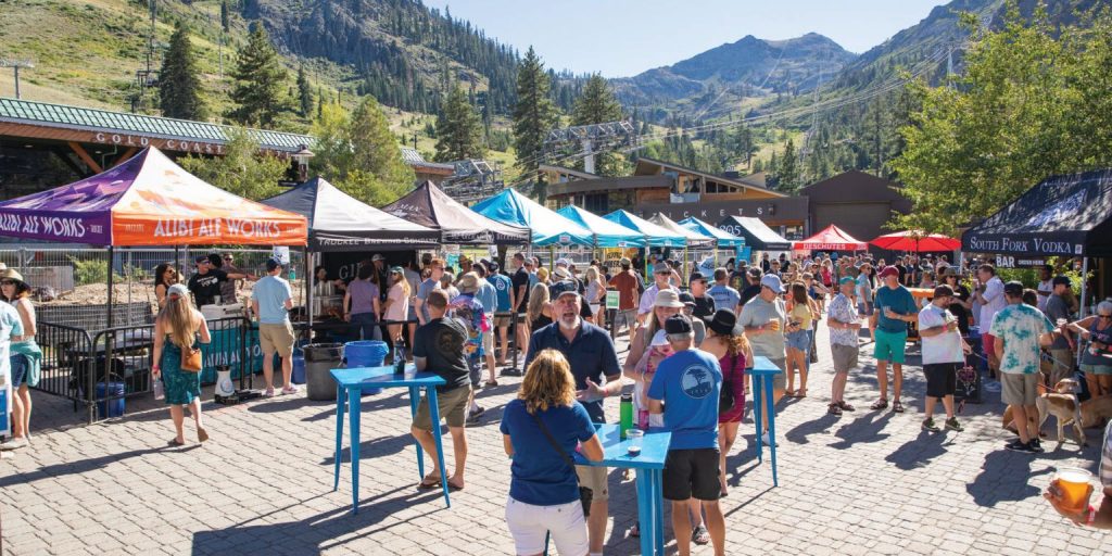 At the base of a mountain, an outdoor festival unfolds with numerous tents and booths. People mingle, drink in hand, while interacting with various vendors. The event is framed by trees and mountainous terrain under a clear blue sky.