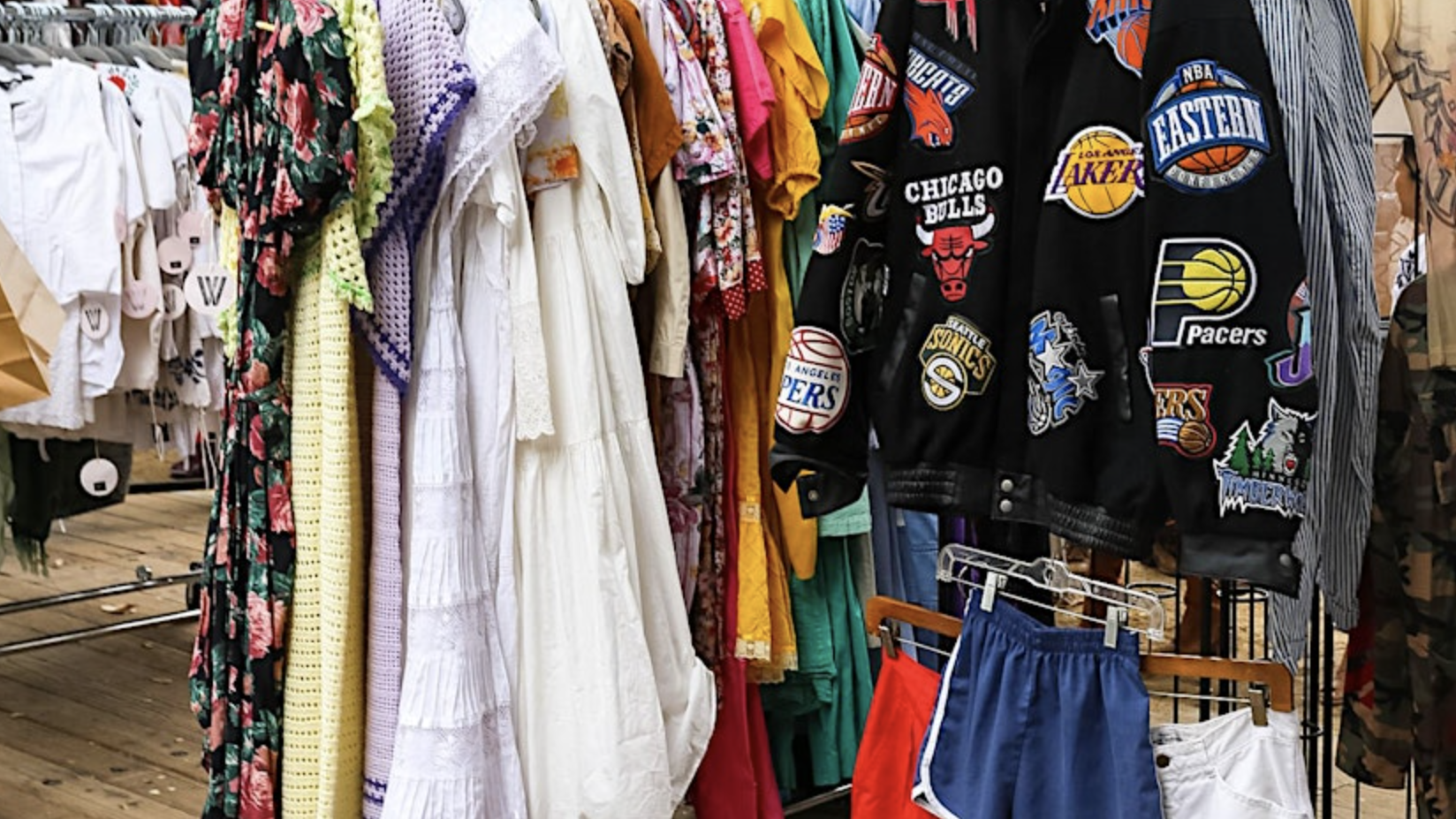 A clothing rack at the Vintage Market features an array of garments. Among them are floral dresses, bright yellow and white outfits, and various patterned tops. A black sports jacket with NBA team logos stands out next to a pair of blue and red shorts. The display captures the nostalgic appeal of yesteryears.