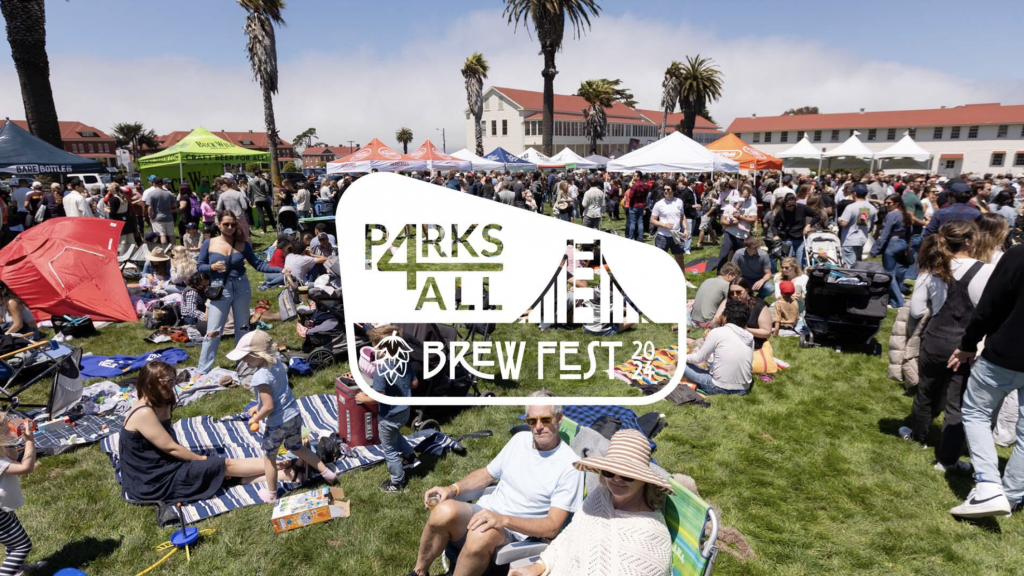 Visitors gather at the Parks4All Brewfest 29, engaging in various outdoor activities. Some attendees relax on blankets and chairs, chatting near the vendor tents. Palm trees and historic buildings form a backdrop under clear skies, enhancing the lively ambiance.