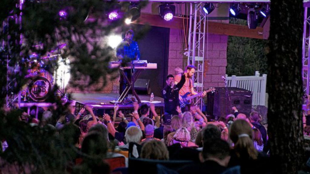 A band performs on an outdoor stage, framed by lit trees and purple lights. Mountains form a distant backdrop. The audience watches closely, some moving their hands to the music's beat. A keyboardist and guitarist are front and center, with a drummer behind them.