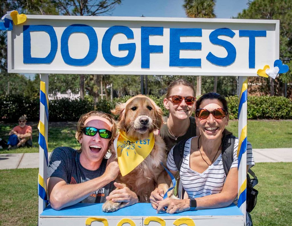 Four people pose cheerfully in a photo booth marked "DOGFEST." They smile, gathered around a Golden Retriever wearing a "Dog Fest" bandana. The summer event takes place on a sunny day, with green grass and clear blue sky as the backdrop. Distant figures and palm trees can be seen in the background.