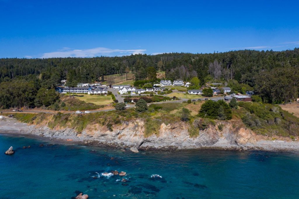 An aerial view captures Little River Inn situated on a tree-covered hill above a rocky shoreline with turquoise waters. The inn includes several white buildings with sloped roofs, all set among green lawns and backed by dense forest.