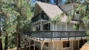 A two-story Breckenridge Chalet stands among tall pine trees. The chalet features a slanted roof and a spacious wrap-around balcony on the upper floor. Dark railings contrast with the white siding, and numerous windows provide views of the forest.