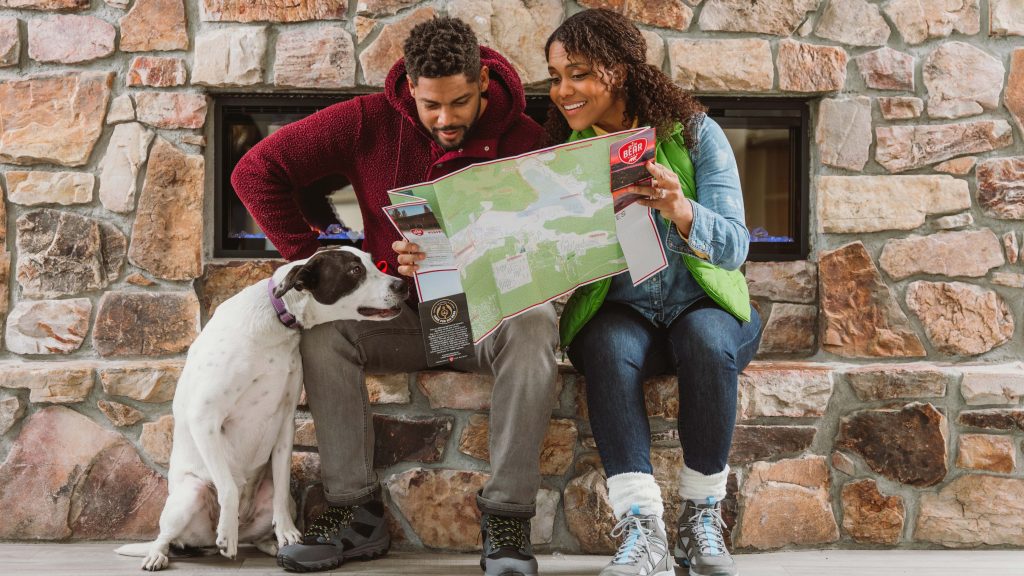 A couple sits on a bench by a stone wall at Big Bear Lake, studying a map. The man wears a burgundy jacket and holds the map. The woman, in a green vest, points to different locations on it. Next to them, their white and black dog sits attentively, eyes fixed on the map.