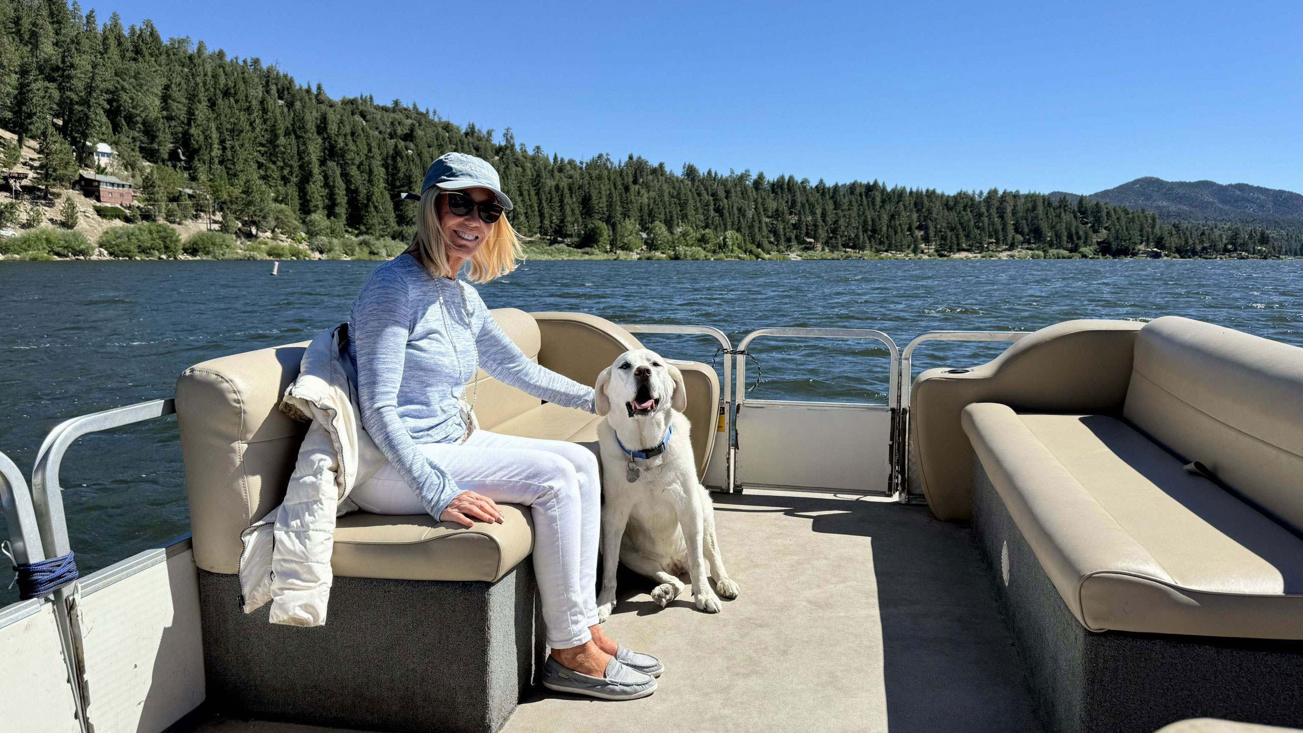 A woman in a light blue outfit and baseball cap sits on a boat beside her white Labrador Retriever. They both enjoy the sunny day on Big Bear Lake, with forested hills and clear skies in the background.