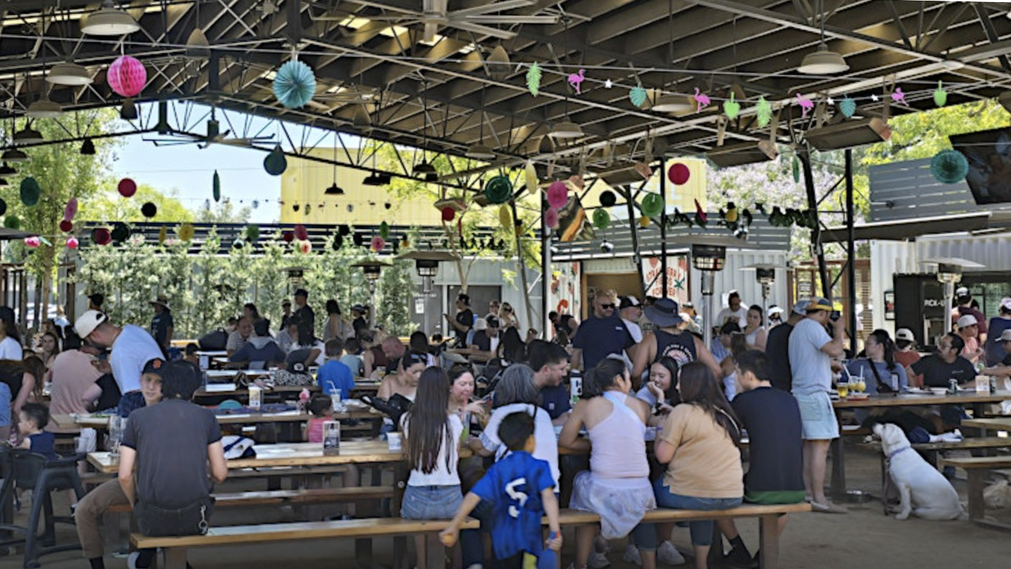 A lively outdoor picnic area, filled with people seated at long tables under a canopy, hosts a 4th of July celebration. Vibrant paper decorations hang from the roof. Adults, children, and dogs enjoy food and drinks against a backdrop of lush greenery.