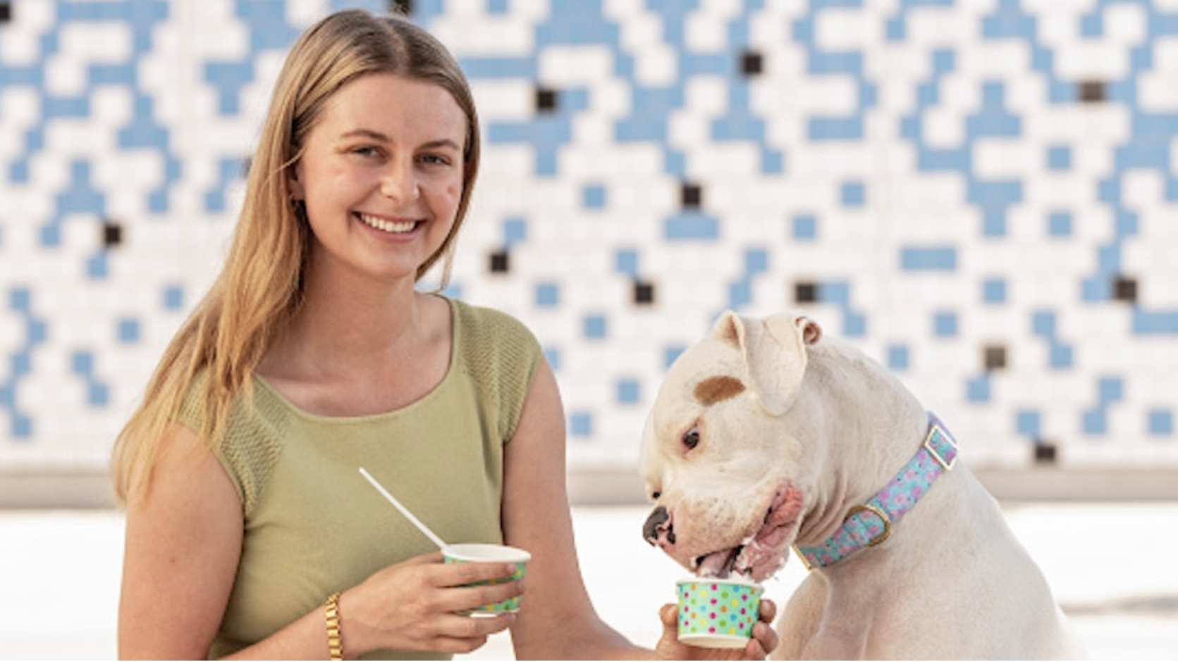 A woman with long blonde hair, dressed in a light green top, smiles as she holds a cup of frozen dessert at an Ice Cream Social. Beside her, a large white dog with a brown patch over one eye licks from an identical cup. The backdrop is a blue and white pixelated wall in West Hollywood.