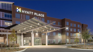 A modern structure in Bakersfield displays a "Hyatt Place" sign illuminated against the night sky. At the entrance, a glass-paneled canopy extends over large windows. Bright lights ensure the area is well-lit, highlighting newly planted trees in a landscaped setting.