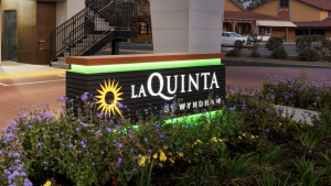 An illuminated sign for La Quinta Inn & Suites by Wyndham Clovis stands out against the night sky. It's surrounded by green and purple flowers. In the background, there is a building with stairs and another structure that has a red roof. The sign features a yellow sun logo and white text on a dark backdrop.